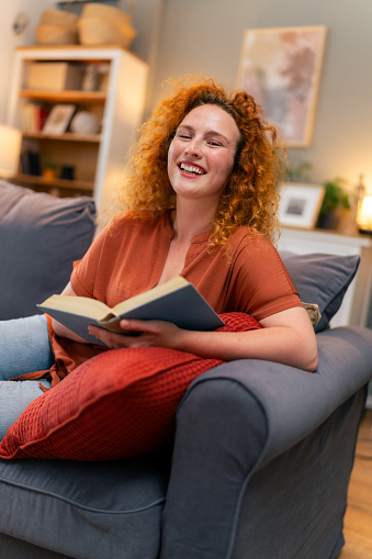 Red-headed woman smiling at camera while holding a book at home.