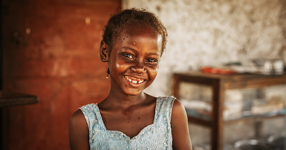 Close Up Portrait of a Shy Authentic African Little Girl Smiling at the Camera with Blurred Background. Black Female Kid Representing Future, Hope, and Acceptance. Documentary Concept