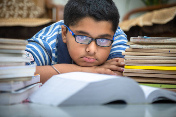 Sad schoolboy looking at the camera with pile of books on the desk. Exhausted little boy learner has tired expression during learning from a lot of books. Consuming education concept