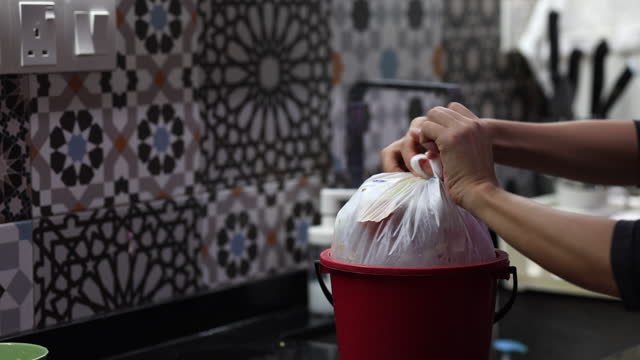 A cropped view of an Asian woman's hands tying a plastic bag containing food waste in the kitchen