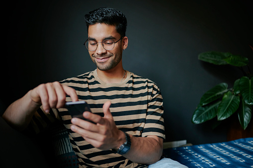 Close-up of smiling young man swiping through a dating app on a smart phone while relaxing on his living room sofa at home