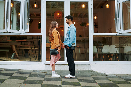 Smiling young couple holding hands together and standing face to face on a sidewalk outside of a cafe