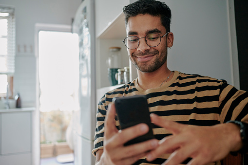 Smiling young man swiping through a dating app on a smart phone while sitting in his kitchen at home