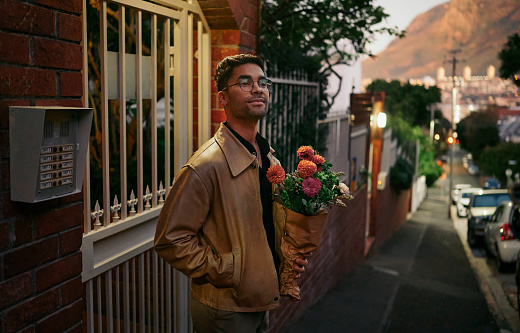 Smiling young man with a bouquet of flowers waiting outside on a sidewalk for his blind date to arrive in the early evening