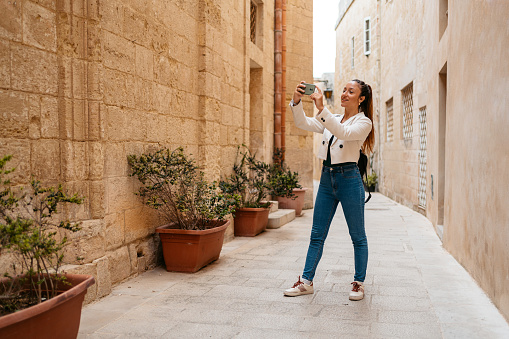 Beautiful young woman taking pictures using her smart phone while standing in an alley in Mdina in Malta.