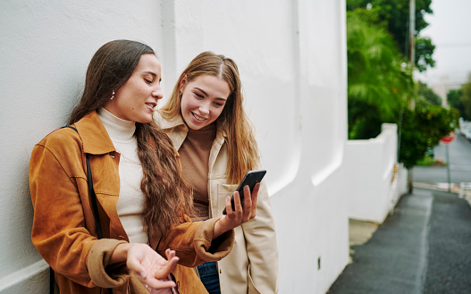 Two smiling young female friends swiping through a dating app on a smart phone while standing outside on a city sidewalk