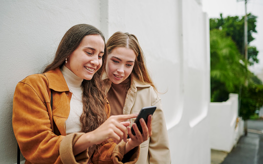 Two smiling young female friends swiping through a dating app on a smart phone while standing together on a city sidewalk