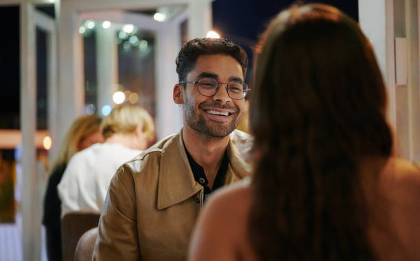 Young man laughing while on a date with a young woman in the evening Young man talking and laughing with a young woman while sitting at a restaurant table during a dinner date blind date stock pictures, royalty-free photos & images