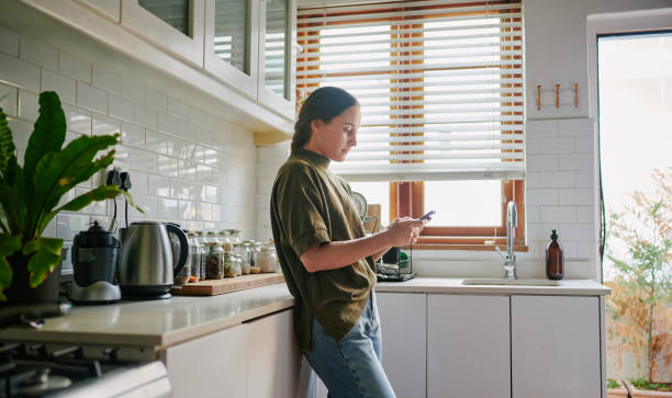 istock Smiling young woman checking her smart phone while standing in her kitchen 1555732915
