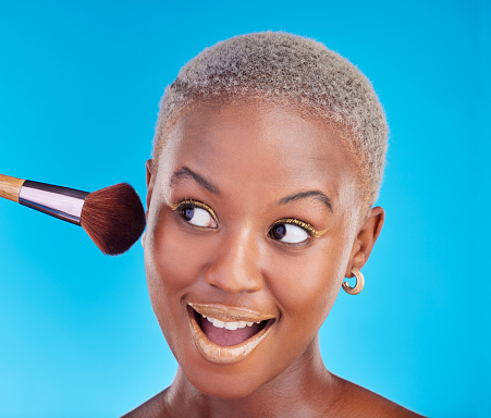 Black woman, face and brush of makeup in studio for application of foundation on blue background. Happy female model, wow and brushing beauty powder on skin with cosmetic tools for facial aesthetic