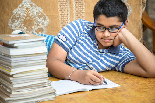 Sad schoolboy looking on a pile of books on the desk. Exhausted little boy learner has tired expression during learning from a lot of books. Consuming education concept