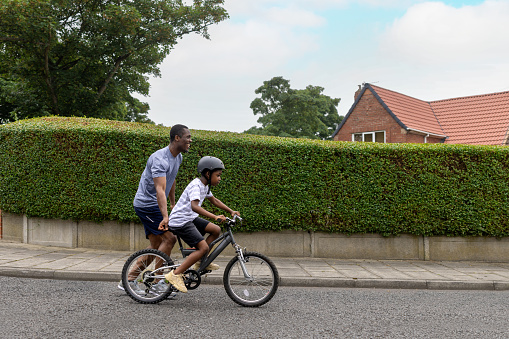 An enthusiastic father teaching his son how to ride a bike in a residential district. The boy ends up riding by himself. They are both wearing casual clothing.