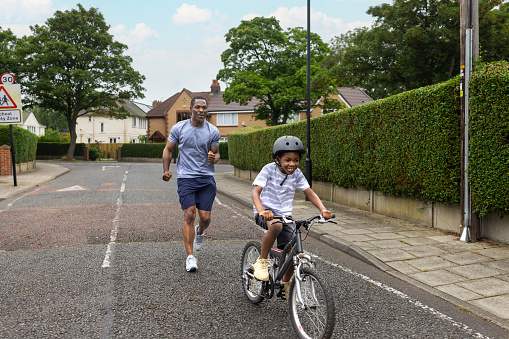 A proud and supportive father teaching his son how to ride a bike on a suburban street where they live. His son is wearing a helmet and is riding by himself.
