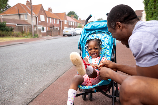A father interacts with his little daughter as she is sitting in her pushchair. He is sharing a positive interaction as he tickles her hand and makes her laugh. They are out on a walk in a rural housing area.