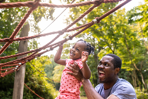 An enthusiastic father lifts up his toddler daughter as she climbs a climbing frame in an outdoor play park. They are both excited and having fun on a bright summer's day.