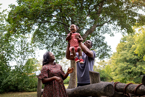 A family walking in the park with their toddler daughter on a summer's day. The father lifts his daughter up in the air to make her giggle for fun. They are surrounded by trees with lush foliage.