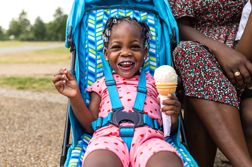 A close-up of a female toddler sitting in her pushchair as she eats an ice cream. She has ice cream all around her mouth as she looks into the camera with a cheeky smile. Her mother is sitting by her side.