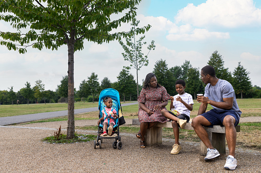 Wide angle perspective of a young family sitting on a bench in their local park on a summer's day.  They are taking a break, eating and drinking. The female toddler is eating ice cream and the young boy is eating grapes and tomatoes, the farther is drinking coffee.