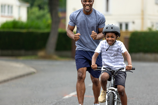 A supportive father cheers his son as he teaches him how to ride a bike in a residential area. The little boy is concentrating as his dad is smiling with excitement. The boy is wearing a helmet and is riding on the road.