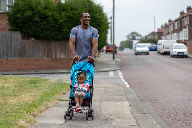 A proud ather walks along a residential footpath in his neighbourhood as he pushes his daughter in her pushchair. He is smiling as he looks into the distance. his daughter is sitting in the pushchair with a serious face.