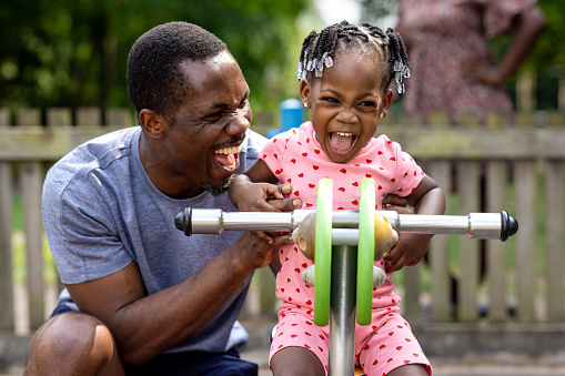 A father and his toddler daughter playing on a spring rocker in their local outdoor play park on a summers day. He is kneeling next to her as he pushes her. They are both pulling excitable facial expressions and having fun. The mother is standing in the background watching.