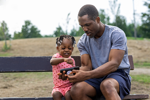 A father and his toddler daughter sitting on a bench in their local park. He is showing his daughter something on his phone as she leans in. She is smiling and enthusiastic.