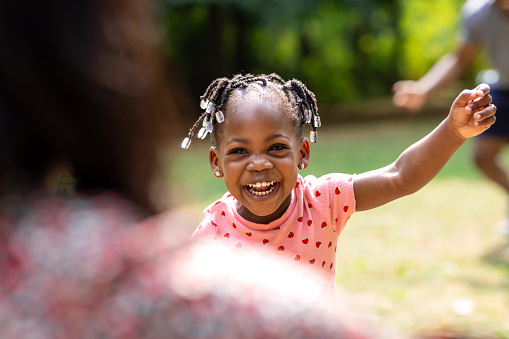 An over-the-shoulder view of  female toddler running towards her mother with open arms on a summers day in their local park. She is smiling with excitment.