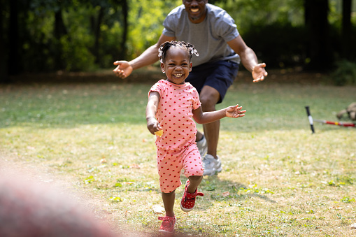 A little girl running away from her father towards her mother at their local park during a sunny summer. The toddler is laughing with excitement as she is running.