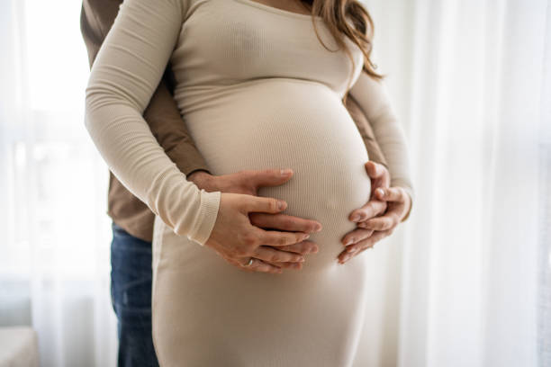 istock Man touching pregnant woman's stomach while standing in room near window 1555809330