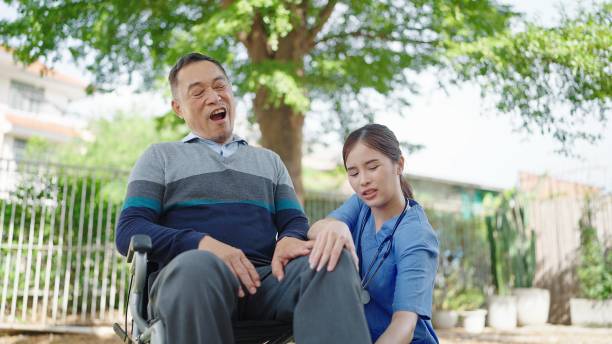 patient asiatique âgé assis dans un fauteuil roulant avec des douleurs aux jambes. l’infirmière soignante prend soin et soutient un patient âgé en fauteuil roulant souffrant de douleurs aux jambes - patient male nurse nurse hospital photos et images de collection