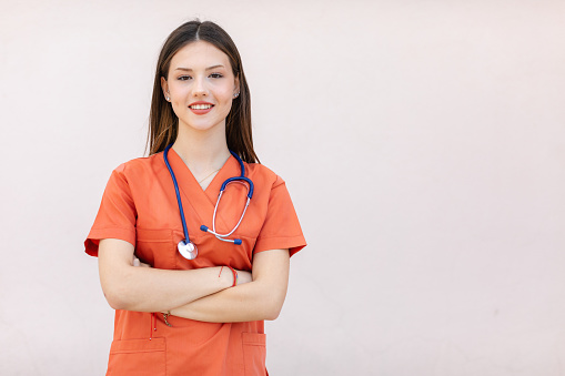 Head and shoulders portrait of cheerful of a paramedic healthcare worker nurse woman with long hair wearing an orange colour surgical uniform and a stethoscope working for EMT, standing, arms folded, smiling, looking at the camera posing in front of a pink background hospital wall.