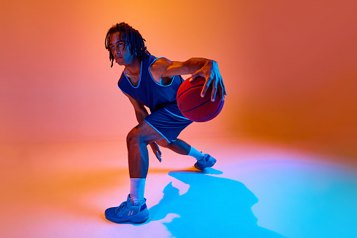 Dynamic image of young competitive male, athlete in motions, training, playing basketball against orange background in neon lights. Concept of professional sport, competition, hobby, game, competition