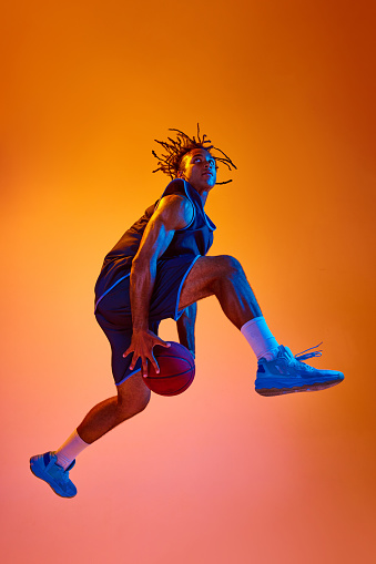Slam dunk position. Athletic young man, basketball player in motion with ball against orange background in neon lights. Concept of professional sport, competition, hobby, game, competition, ad