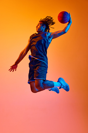 Full-length image of young sportsman, basketball player in motion, jumping with ball against orange background in neon lights. Concept of professional sport, competition, hobby, game, competition, ad