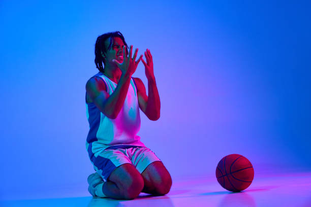 istock Loosing game. Young guy, basketball player sitting on knees in sadness against gradient blue background in neon lights 1555862062