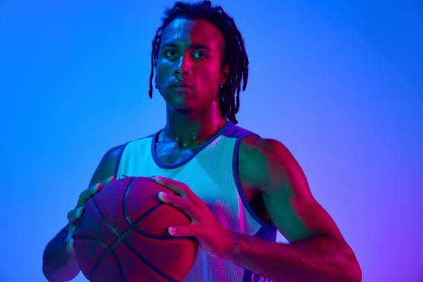 istock Young man with dreads, basketball player in uniforma posing with ball against gradient blue background in neon lights 1555862591