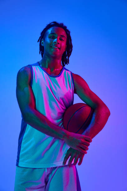 istock Young man with dreads, basketball player in uniforma posing with ball against gradient blue background in neon lights 1555862594