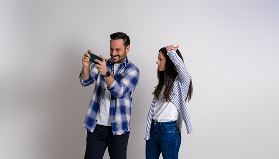 Girlfriend with head in hand staring furiously at excited boyfriend playing video game on smart phone against background. Serious woman looking at cheerful man using cellphone