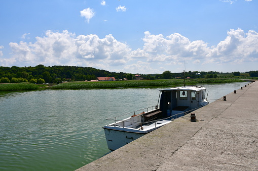 A close up on a boat or other passenger vessel traversing a vast and deep ocean or sea seen in some distance to the coast covered with forests and moors spotted on a sunny summer day in Poland