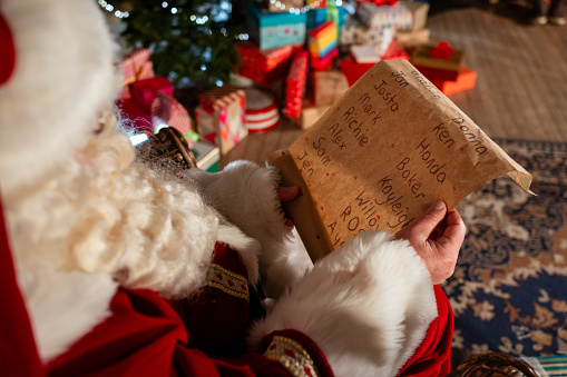 Over the shoulder view of a man dressed as Santa reading a list titled 'naughty or nice' surrounded by presents. There is a Christmas tree and the scene is set up as a Santas grotto, the actor waiting to meet children.