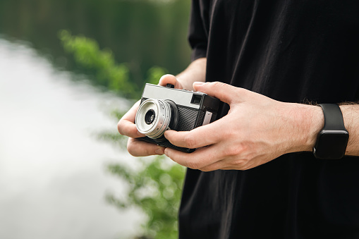 Old film camera in a male hand on a blurred background of the river, the concept of travel, tourism and analog photography.