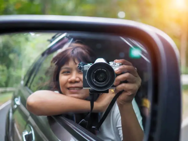 Photo of A woman holds a digital camera and takes a picture of herself smiling reflected in the car mirror.