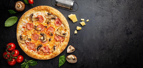 Tasty mushrooms and ham pizza and cooking ingredients tomatoes and basil on dark background. Top view