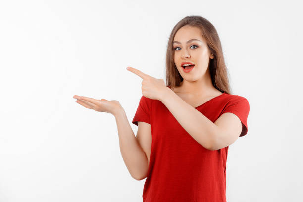 istock Surprised lovely girl points left at promo banner, shows advertisement on blank white space. Young brunette female looking amazed by promotion offer, stands over white background 1555948150