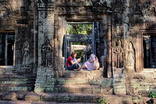 Siem Reap, Cambodia, December 19, 2018. Two Asian girls are sitting on the threshold of an ancient stone structure.