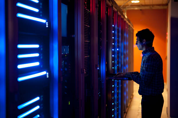 IT Engineer in Action Configuring Servers The interior of a modern server room in a data center.  An IT engineer is busy configuring the servers. The room is dark, but the servers themselves are lit.  The servers at the left side are lit in blue, while the ones at the other end are lit by an orange light source coming from the right side. data cable stock pictures, royalty-free photos & images