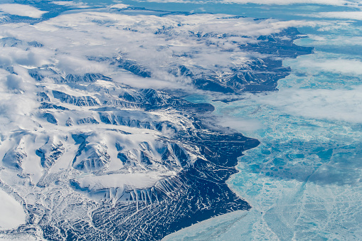Aerial view from 38,000 feet of the mountains and hills on the coastline of Greenland in the Arctic circle mostly covered in snow and low clouds hanging on the ground; sea covered with ice