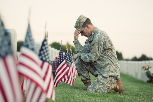 A soldier in ACU fatigues kneels in front of a grave with several American flags in front of it.  There is bright green grass under all, and another row of markers is behind the soldier, with flowers in front of them.