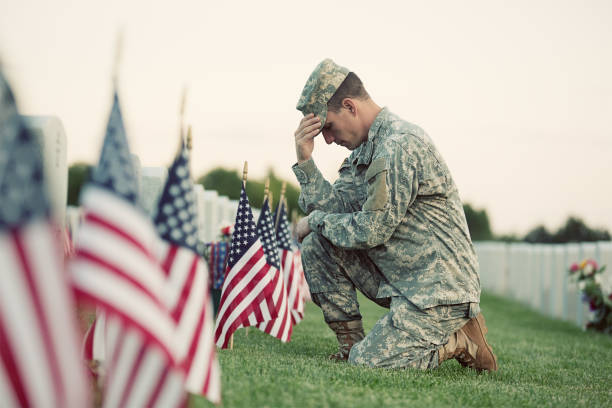 Soldier kneeling at grave A soldier in ACU fatigues kneels in front of a grave with several American flags in front of it. There is bright green grass under all, and another row of markers is behind the soldier, with flowers in front of them. soldier kneeling at grave stock pictures, royalty-free photos & images