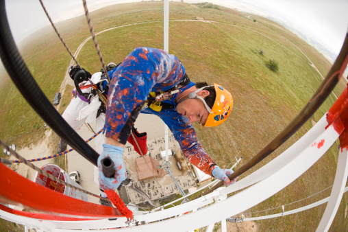 Man high above ground using rope access saftey equipment to paint radio tower.  Wide angle view looking down for scary perspective.